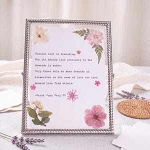 May include: A rectangular silver metal frame with a twisted rope border containing a white paper sheet with the printed text: Genuine love is demanding. But its beauty lies precisely in the demands it makes. Only those able to make demands on themselves in the name of love can then demand love from others. -Saint John Paul II. The paper is decorated with various pressed pink and white flowers and green leaves.