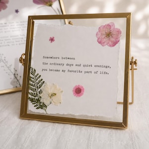 May include: A square gold metal frame containing a piece of white paper with torn edges. The text reads: Somewhere between the ordinary days and quiet evenings, you became my favourite part of life. Pressed pink and white flowers and a green fern leaf.