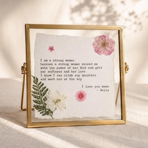 May include: A square gold-toned metal floating frame containing a piece of textured white paper with deckled edges. The paper features black typewriter-style text about a strong mother and is decorated with pressed pink flowers and a green fern.