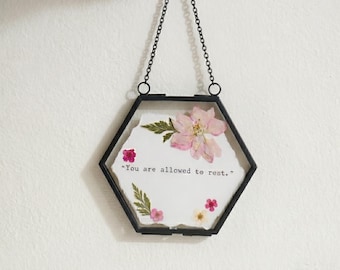 Puede incluir: Un marco hexagonal de metal negro que cuelga de una cadena negra, con un inserto de papel blanco con flores prensadas rosas y blancas y hojas verdes. El texto "You are allowed to rest." está impreso en una fuente negra estilo máquina de escribir.