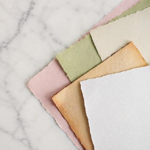 May include: A collection of five rectangular sheets of handmade paper with rough, deckled edges. The stack includes colors of soft pink, sage green, cream, tan, and bright white, each featuring a textured, fibrous surface finish.