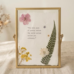 May include: A rectangular gold metal frame containing a piece of off-white textured paper with pressed flowers and a green fern leaf. The paper features the printed text: "You were made of poetry before the world called called you ordinary."