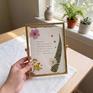 May include: A rectangular brass-toned metal frame containing a piece of textured white paper with deckled edges. The paper features black printed text that reads: Some souls never truly leave us. They remain in quiet corners, old routines, and the love that still lives inside us. Pressed flowers in pink, yellow, and white, along with a green fern leaf, are arranged around the text.