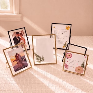 May include: A collection of four rectangular floating picture frames with thin gold and black metal borders. The frames display photographs of a couple, a pressed green fern leaf, and printed text with pressed flowers on white paper backgrounds.