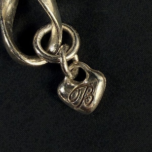Puede incluir: Una pulsera de eslabones de metal plateado con un peque&ntilde;o dije en forma de coraz&oacute;n que cuelga de una argolla circular. El dije tiene grabada una letra B estilizada en cursiva en su parte frontal, con un acabado pulido que refleja la luz.
