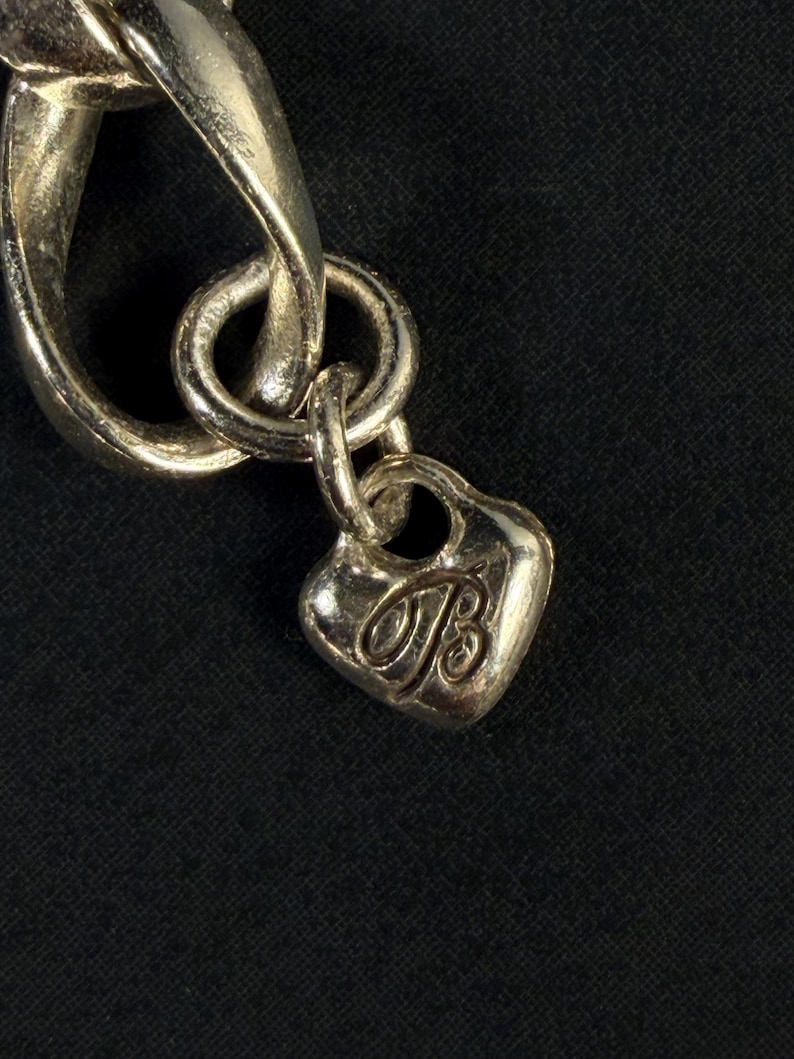 Puede incluir: Una pulsera de eslabones de metal plateado con un peque&ntilde;o dije en forma de coraz&oacute;n que cuelga de una argolla circular. El dije tiene grabada una letra B estilizada en cursiva en su parte frontal, con un acabado pulido que refleja la luz.