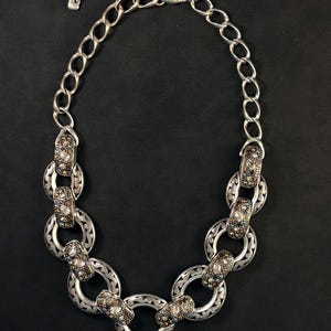 Puede incluir: Un collar de cadena en tono plateado que presenta una serie de eslabones circulares entrelazados. Cada eslab&oacute;n est&aacute; decorado con intrincados patrones de volutas y acentuado con peque&ntilde;as piedras incrustadas multicolores en tonos marr&oacute;n, azul y transparente.