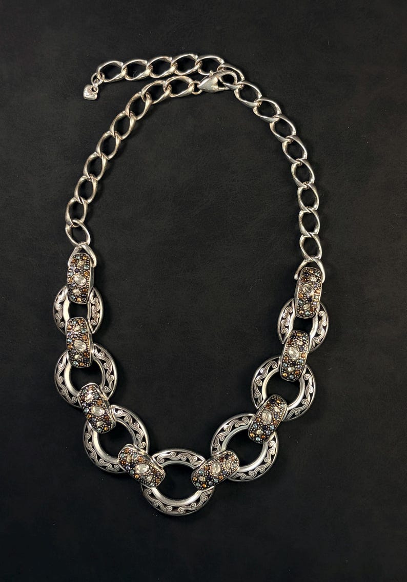 Puede incluir: Un collar de cadena en tono plateado que presenta una serie de eslabones circulares entrelazados. Cada eslab&oacute;n est&aacute; decorado con intrincados patrones de volutas y acentuado con peque&ntilde;as piedras incrustadas multicolores en tonos marr&oacute;n, azul y transparente.