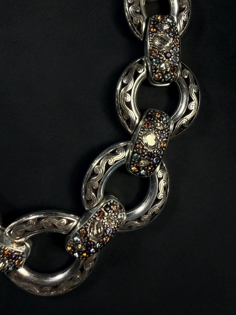 Puede incluir: Un collar de cadena de plata con eslabones circulares alternos con grabados de volutas intrincados y eslabones rectangulares incrustados con peque&ntilde;as piedras preciosas multicolores en tonos &aacute;mbar, azul y transparente.
