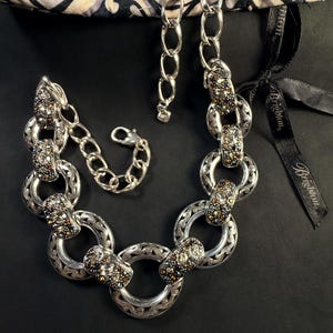 Puede incluir: Un collar de eslabones en tono plateado que presenta anillos circulares alternos con intrincados grabados y segmentos rectangulares incrustados con peque&ntilde;os cristales multicolores. El collar incluye un cierre de langosta y una cadena extensora.