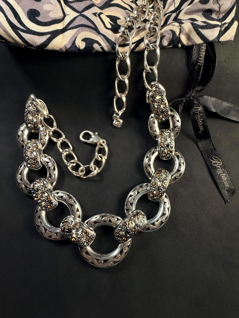 Puede incluir: Un collar de eslabones en tono plateado que presenta anillos circulares alternos con intrincados grabados y segmentos rectangulares incrustados con peque&ntilde;os cristales multicolores. El collar incluye un cierre de langosta y una cadena extensora.