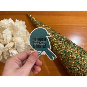 May include: A teal bird-shaped sticker with the text "SHE NEEDS ME I have to remind her to breathe TO FIND HER." The sticker is held in front of a gold sequined cone and a bouquet of white flowers.