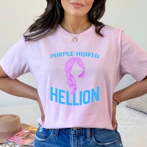 Puede incluir: Camiseta rosa claro con la frase "PURPLE-HAIRED HELLION" en azul y una ilustración morada de una trenza. La camiseta es de material suave, ideal para uso diario.