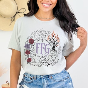 May include: A light gray short-sleeve t-shirt featuring a circular graphic design. The print includes a dragon, a fox, a deer, roses, and a flaming tree surrounding the letters FFG in a central frame. The design uses black, red, and purple.