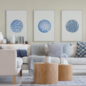 May include: A modern living room with a white sofa, grey throws, and patterned cushions. Three framed artworks with blue circular patterns are displayed. Two wooden side tables and decorative items add to the contemporary design.