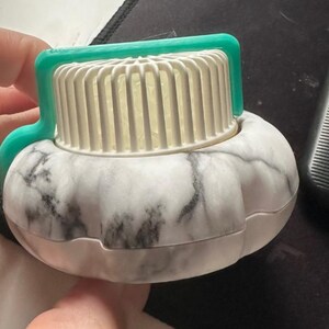 May include: A white and gray marble-patterned object with a white, ridged top and a teal handle. The object is round with a ridged top and a teal handle. The object is likely a beauty or skincare tool.