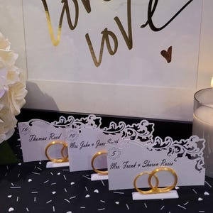 May include: Wedding place cards with gold ring accents. Each card features a name and table number. The cards are displayed on a black and white patterned surface, with a lit candle and a bouquet of white flowers in the background.