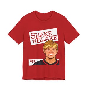 Op de afbeelding: Rood t-shirt met de tekst "SHAKE 'N BLAKE" in het wit op een witte rechthoek. Daaronder een cartoon illustratie van een lachende persoon met blond haar. Het nummer "53" wordt eronder weergegeven.