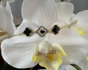 Puede incluir: Una pulsera dorada con tres colgantes en forma de trébol. Dos colgantes son negros, y el colgante central es blanco con un diseño floral negro. La pulsera está colocada sobre un fondo floral blanco y amarillo.