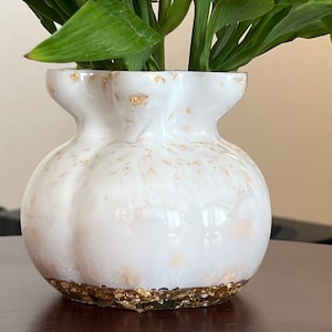 Op de afbeelding: Een witte, ronde vaas met meerdere lobben en een geschulpte rand. Het oppervlak is voorzien van verspreide bladgoudvlokken, met een dichte band van bladgoud rond de basis. Groene stengels en witte bloemen zijn zichtbaar in de vaas.