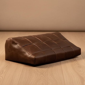 Puede incluir: Un otomán de cuero marrón con un diseño tejido. El otomán rectangular es bajo y tiene una parte superior ligeramente inclinada. El cuero es de un color marrón oscuro intenso y las costuras son visibles.