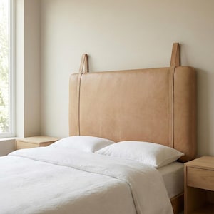 May include: A light brown leather headboard with hanging straps, paired with white bedding. The headboard is rectangular and has a padded appearance. Two matching wooden nightstands are on either side of the bed.