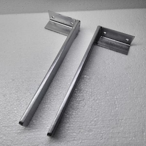 Puede incluir: Dos soportes para estantes de metal en forma de L con acabado plateado. Cada soporte presenta un brazo vertical largo y plano, y una placa de montaje horizontal más corta en la parte superior con dos orificios circulares preperforados.