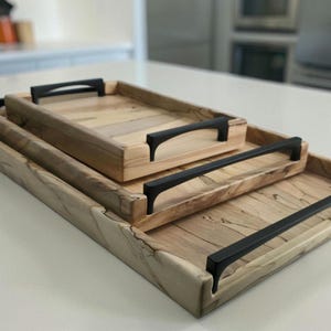 May include: A set of three wooden serving trays with black handles. The trays are rectangular with a light wood grain pattern. The handles are curved and attached to the sides of the trays. The trays are nested, with the largest tray at the bottom.