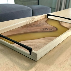May include: Wooden serving tray with a natural wood grain and a yellow resin river design. The tray has black handles and is rectangular. A decorative piece for serving or display.