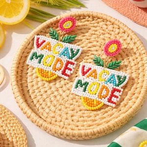 May include: Colorful beaded earrings with the words "VACAY MODE". The earrings feature a white base with the text in red, yellow, green, and blue beads. They are adorned with a pink flower and green leaves.