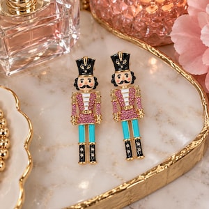 May include: A pair of nutcracker earrings featuring black hats, pink crystal-encrusted jackets, bright turquoise trousers, and black boots with gold-tone metal accents. The figures have painted faces with mustaches and are finished with clear crystals.