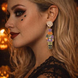 May include: A pair of Halloween-themed earrings. The earrings feature a gold-colored metal design with a pearl cluster at the top. The dangling charm is a nutcracker figure with a pumpkin head, wearing a purple coat and holding a black cat.