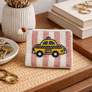 May include: A rectangular coin purse featuring a beaded surface with vertical white and pink stripes. The center displays a beaded yellow taxi cab graphic with blue windows, black wheels, and the letters NYC printed on the side. It has a white zipper.