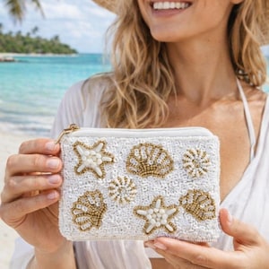 May include: White beaded clutch with gold and white seashell designs. The clutch has a gold zipper and is held by a person on a beach. The bag features various seashell and starfish patterns.