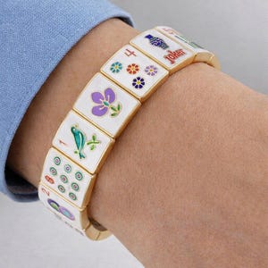 May include: A gold-toned bracelet featuring rectangular white tiles with colorful Mahjong symbols. The tiles display various designs, including flowers, dots, and a fish, with a 'Joker' tile visible. The bracelet is worn on a wrist.