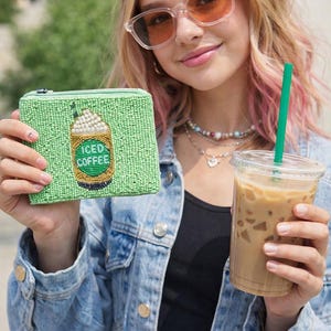 May include: A green beaded coin purse with an "ICED COFFEE" design, held next to a clear plastic cup of iced coffee with a green straw. The purse has a zipper closure and a coffee cup design.