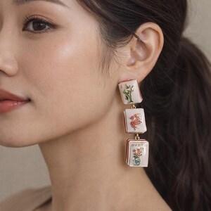 May include: Dangling earrings with three rectangular, off-white pendants. Each pendant features a different floral design in red, green, and yellow. The earrings have a gold-colored metal frame and are connected by small gold chains.