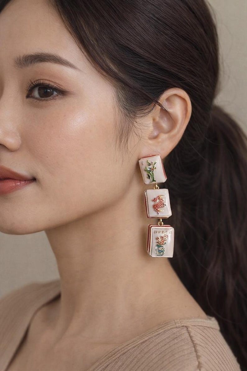 May include: Dangling earrings with three rectangular, off-white pendants. Each pendant features a different floral design in red, green, and yellow. The earrings have a gold-colored metal frame and are connected by small gold chains.