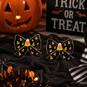 May include: Two black enamel bow-shaped pins featuring a repeating pattern of small candy corn pieces. Each bow has a larger, central candy corn design at the knot. The pins have a glossy finish with gold-toned metal edges and a secure backing.