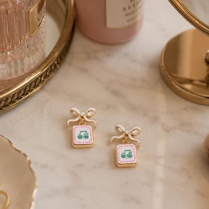 May include: A pair of gold-toned earrings with a white bow and a square pendant. The pendant features a pink background with a green golf cart design. The earrings are set on a marble surface with other beauty accessories.