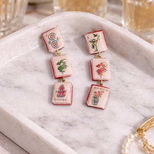 May include: A pair of dangle earrings featuring a series of rectangular charms with red borders and gold-colored connectors. Each charm displays a different colorful illustration, including flowers, a dragon, and the word "Joker". The earrings rest on a marble tray.