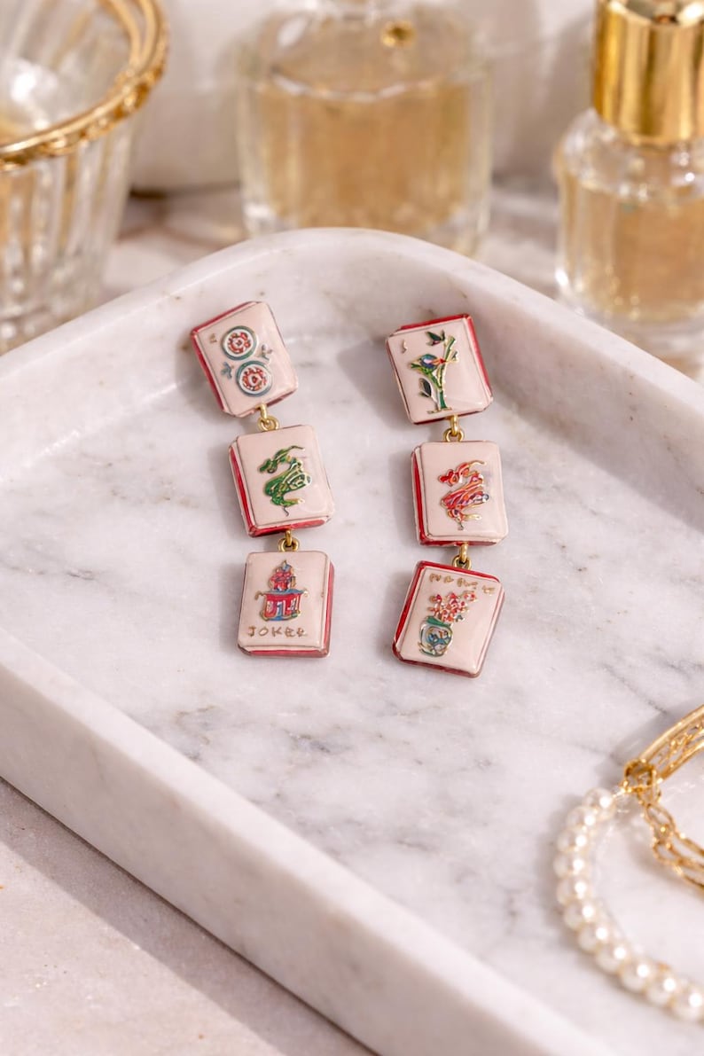 May include: A pair of dangle earrings featuring a series of rectangular charms with red borders and gold-colored connectors. Each charm displays a different colorful illustration, including flowers, a dragon, and the word "Joker". The earrings rest on a marble tray.