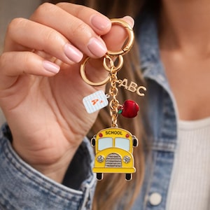 May include: A gold-colored keychain with school-themed charms. The charms include a yellow school bus, a red apple, a white A+ report card, and ABC letters. The keychain is held by a person.