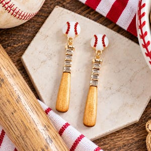 May include: A pair of drop earrings featuring a white baseball charm with red crystal stitching attached to a gold-tone baseball bat charm. The bat handle is embellished with a row of clear rectangular crystals, while the barrel is a light brown wood.