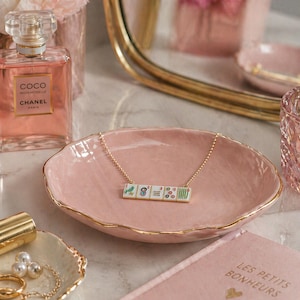 May include: A pink ceramic jewelry dish with a gold rim holds a necklace with a pendant of small, decorative tiles. A bottle of perfume and a small tray with pearls are also visible.