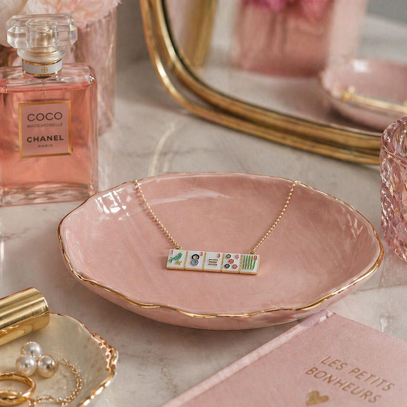 May include: A pink ceramic jewelry dish with a gold rim holds a necklace with a pendant of small, decorative tiles. A bottle of perfume and a small tray with pearls are also visible.