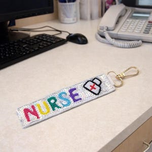 May include: A beaded keychain with the word "NURSE" in colorful letters. The keychain has a nurse's hat icon with a red cross. The background is a light-colored surface with office equipment.