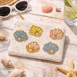 May include: A white beaded clutch with a gold zipper, adorned with colorful seashell designs. The clutch is surrounded by seashells, sunglasses, a straw hat, and a drink with a lime slice, suggesting a beach theme.