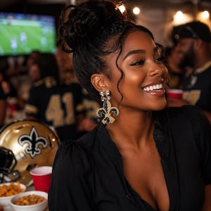 May include: A smiling person wearing black and gold fleur-de-lis earrings. The earrings are beaded and feature a black fleur-de-lis design with gold accents. A gold football helmet with a fleur-de-lis logo is visible in the background.