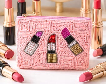 May include: A pink beaded cosmetic bag with three lipstick designs. The bag is surrounded by various lipsticks in gold and black tubes. A gold-framed mirror is in the background.